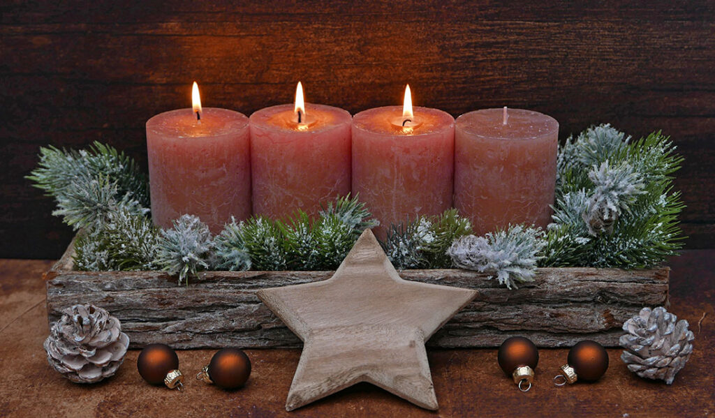 Vier Kerzen brennen in einem weihnachtlichen Gesteck mit Tannenzweigen, Holzstern und winterlicher Dekoration.